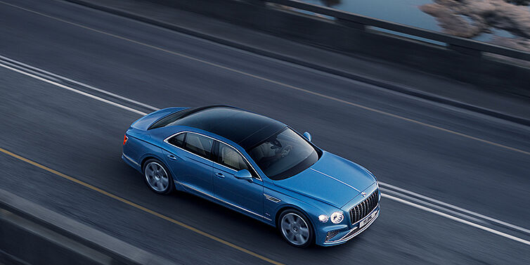 Bentley Paris Seine New Bentley Flying Spur Azure sedan in Light windsor blue paint by Mulliner, with 22 inch Azure wheels seen from overhead driving dynamically on a country road