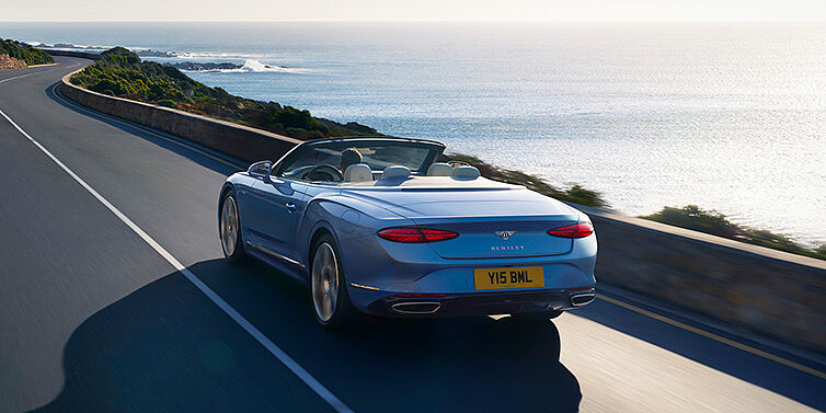 Bentley Paris Seine New Bentley Continental GTC Azure convertible rear view in Light Windsor blue paint with 22 inch ten spoke wheels driving dynamically on an ocean road in the sunshine